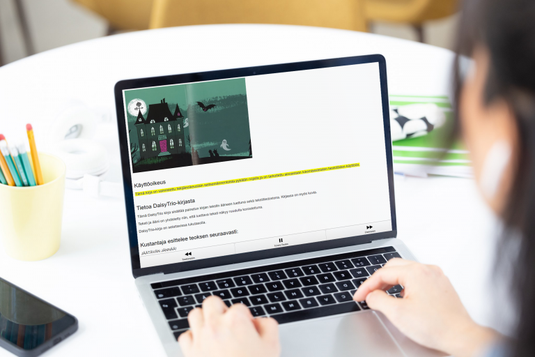 Henkilö tietokoneella takaa viistosti kuvattuna. Tietokoneen ruudulla näkyy DaisyTrio Yökoulu-yhdistelmäkirja. Vieressä pöydällä kyniä kynätelineessä ja puhelin.