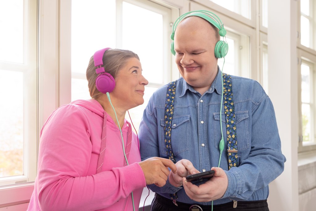Två personer står sida vid sida framför fönstret och ser leende in i varandras ögon. Båda har hörlurar på sig, den andra håller i en mobiltelefon som de båda pekar på. Bild: Tillgänglighetsbiblioteket Celia.