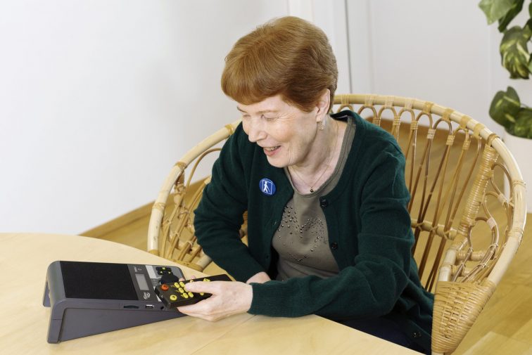 En person sitter vid ett bord, med en svart spelare för böcker på bordet framför sig. Hen har en fjärrkontroll i handen.