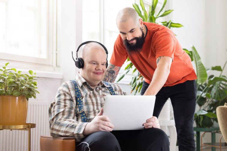 En person sitter i en stol framför fönstret med en bärbar dator öppen i knät och hörlurar i öronen. En annan person står bredvid hen, böjd framåt för att titta på datorskärmen. I bakgrunden finns gröna växter.
