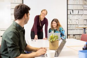 Tre leende personer sitter runt ett stort konferensbord. Två av dem sitter ner med bärbara datorer framför sig, medan en står upp. På bordet finns en grön krukväxt.